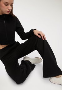 Czarna, krótka bluza z zamkiem i kieszenią z przodu, zestawiona z szerokimi czarnymi spodniami dresowymi. Model nosi białe buty sportowe.