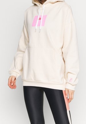 Woman wearing an oversized cream hoodie with a pink square logo and black leggings with pink and white stripes.