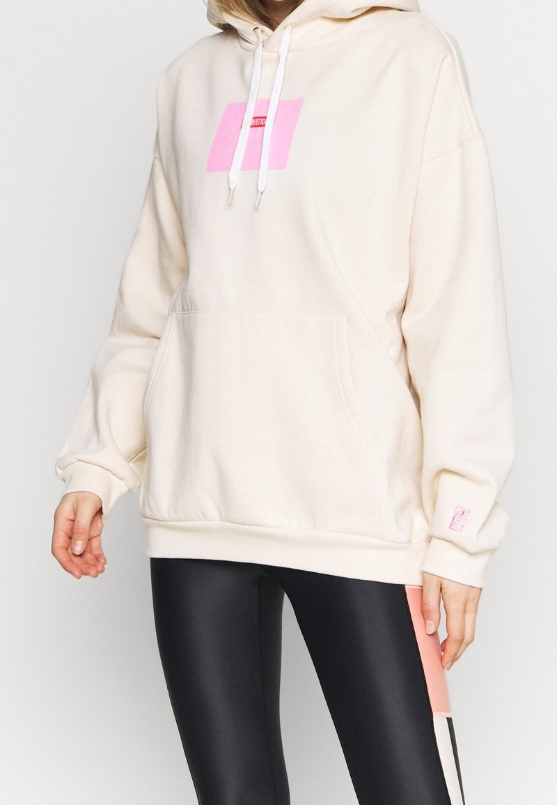 Femme portant un sweat à capuche crème oversize avec un logo carré rose et un legging noir avec des rayures roses et blanches.