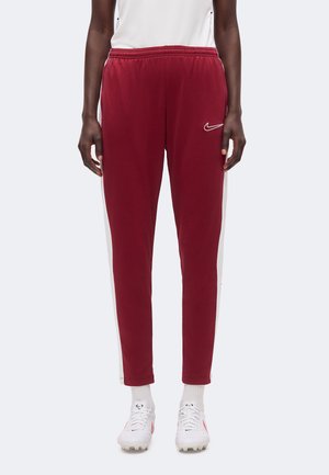 Personne portant un pantalon de sport bordeaux avec des bandes blanches sur les côtés, un haut blanc, des chaussettes blanches et des crampons blancs, debout devant un fond uni.