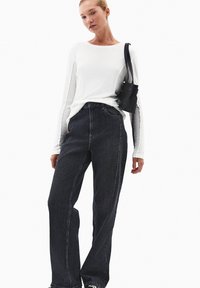 Biała koszula z długim rękawem, czarne jeansy z wysokim stanem, oba wykonane z materiału o gładkiej teksturze. Mała czarna torebka przewieszona przez ramię.