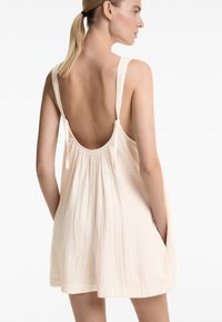 Hellbeige, ärmelloses Kleid mit tiefem Rückenausschnitt, dünnen Trägern und seitlichen Taschen. Hergestellt aus strukturiertem, atmungsaktivem Stoff. Kurze Passform.