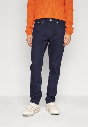 Jeans azul oscuro con corte recto, cinco bolsillos y herrajes de metal. Combinados con un suéter de punto naranja y zapatillas beige.