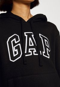 Sweat à capuche noir en matière douce avec une poche kangourou. Dispose d'un grand logo "GAP" brodé en blanc avec un contour noir.
