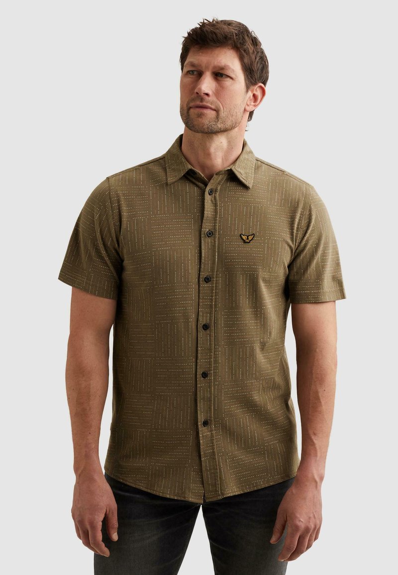 Homme portant une chemise à manches courtes vert olive à motifs, avec des boutons noirs et un petit logo brodé sur la poitrine gauche.