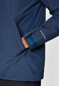 Veste imperméable navy avec une poche zippée, des fermetures par velcro aux poignets et une texture légère et lisse. Affichant le logo "DRYVENT".