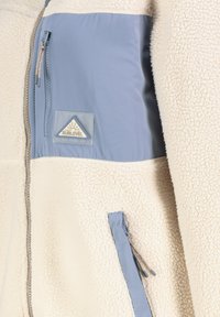 Fleece-Jacke mit weißer Textur, blauen Stoffpaneelen, Fronttaschen mit Reißverschluss und einem dreieckigen Logopatch auf der Brust.