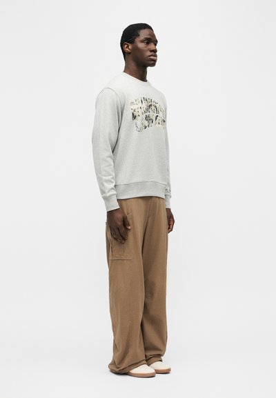 Ung mann står vendt mot høyre iført en lysegrå grafisk genser, løse brune bukser med sidelomme og hvite slip-on sko.