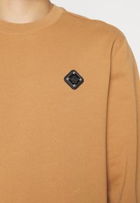 Hnědá Mikina se žebrovaným kulatým výstřihem, s diamantovým černým patchem se stříbrnými akcenty na levé části hrudi. Materiál ze směsi bavlny.