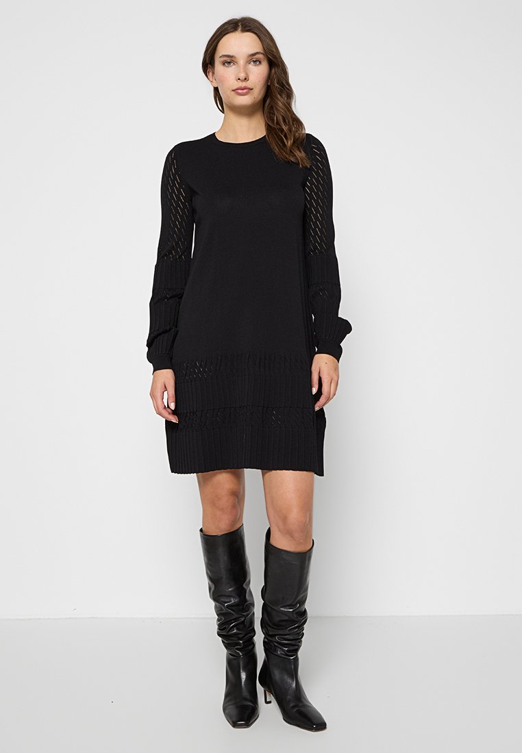 Femme se tenant debout, portant une robe tricotée noire à manches longues avec des manches et un ourlet à motifs, assortie à des bottes en cuir noires montant jusqu'aux genoux.