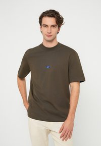 Olivengrønn kortermet T-skjorte i bomullsstoff, med rund hals og en liten blå logopatch på brystet.