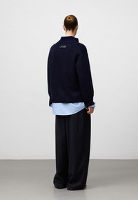 Maglione navy con colletto alto e orlo a costine, indossato sopra una camicia azzurra chiara. Dettaglio della etichetta nera sul retro. Silhouette a vestibilità ampia.