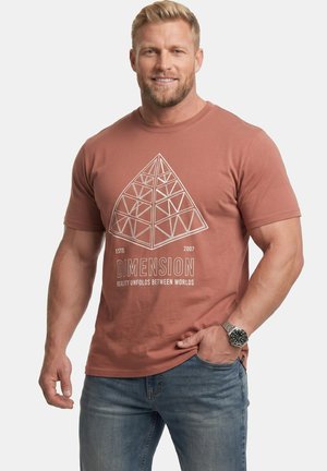 Muskularny mężczyzna noszący rdzawą koszulkę z geometrycznym wzorem piramidy i tekstem "Dimension: Reality unfolds between worlds" oraz niebieskie dżinsy.
