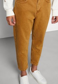 Pantaloni in velluto a coste marroni con una vestibilità affusolata, dotati di tasche frontali e cuciture in vista, abbinati a sneaker bianche.