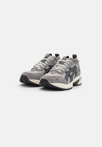 Grijze sportschoenen met een combinatie van mesh en suède materialen, een zwarte accentstreep, witte veters en een gestructureerde rubberen loopzool.