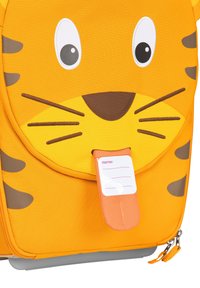 Orange backpack features a tiger face design with white eyes, brown accents, and a name tag holder. Made of durable fabric with a zip closure.