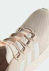 Zapato deportivo beige con tejido texturizado, cordones elásticos y tres rayas blancas en el lateral. Punta redondeada y detalles tonales sutiles.