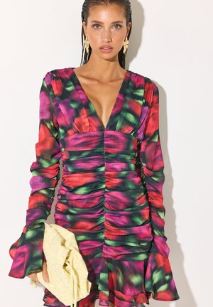 Femme portant une robe longue froncée à manches longues avec un décolleté en V profond et un motif multicolore rouge, vert et violet, tenant une pochette texturée jaune clair.