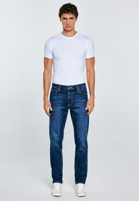 Jeans de mezclilla azul para hombre con corte recto, diseño de cinco bolsillos y sutil textura desgastada; combinados con una camiseta blanca simple y zapatillas blancas.