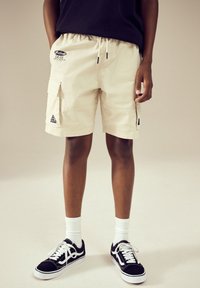 Beige cargo shorts with elastic waistband, two side pockets, and logo detail on the left thigh. Paired with black sneakers and white socks.
