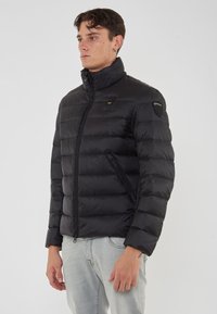 Uomo che indossa una giacca trapuntata nera con colletto alto e jeans azzurri, in posa davanti a uno sfondo bianco neutro.