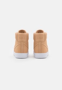 Un paio di sneakers Nike high-top color sabbia viste da dietro con suole bianche e logo Nike sulla linguetta del tallone, su uno sfondo bianco.