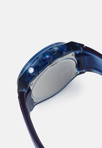 Transparante blauwe plastic horloge met een ronde wijzerplaat, zilveren achterkant en drie blauwe knoppen aan de zijkant; heeft een flexibele donkerblauwe band.