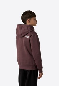 Lilla hettegenser med stor hette, ribbestrikkede mansjetter, rett kant og en hvit logo på øvre del av ryggen. Brukes med svarte bukser.