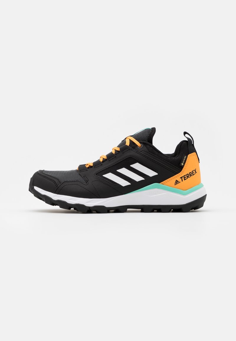 Chaussure de sport noire avec un dessus texturé, des lacets orange et un logo blanc à trois bandes. Présente un accent vert et blanc sur la semelle.