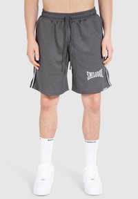 Pantalones cortos deportivos grises con textura acanalada, una cintura elástica, un logo blanco y franjas laterales en contraste. Combinados con zapatillas blancas y calcetines.