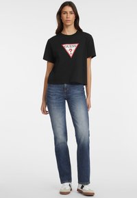 T-shirt noir court avec un logo triangulaire rouge sur lequel est inscrit « GUESS ? » en lettres blanches ; associé à un jean droit bleu et des baskets blanches.