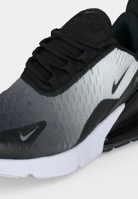 Κοντινό πλάνο μαύρου και γκρι παπουτσιού Nike Air Max 270 που αναδεικνύει τα κορδόνια, το άνω μέρος από διχτυωτό υλικό και τη λευκή ενδιάμεση σόλα με ορατό αερόσακο στη φτέρνα.
