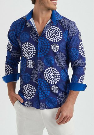 Chemise bleue à motifs avec divers designs circulaires, fabriquée en tissu lisse, avec un col à boutons et des poignets bleus contrastants.