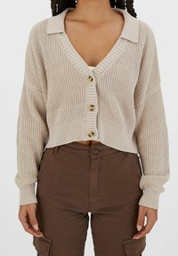 Beige stickad cardigan med V-ringning, knappstängning och ribbad mudd. Korta ärmar, strukturerat material och subtila bruna knappar.