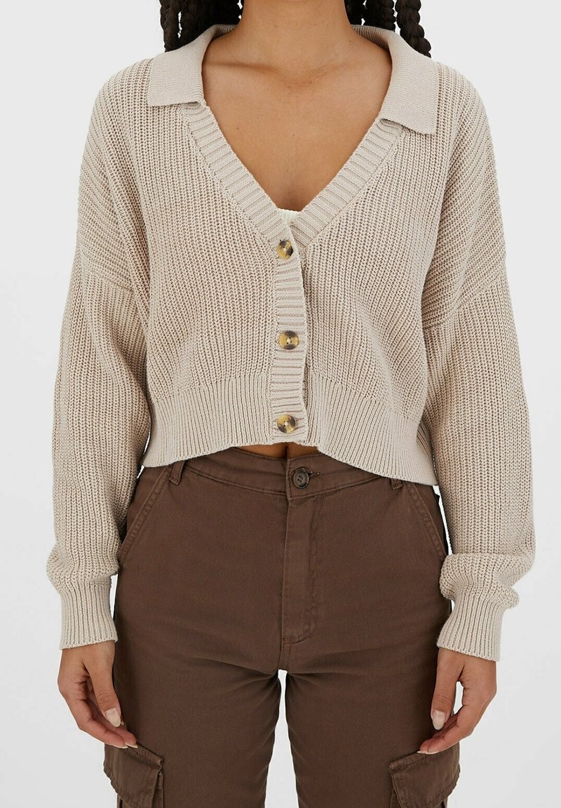 Beige stickad cardigan med V-ringning, knappstängning och ribbad mudd. Korta ärmar, strukturerat material och subtila bruna knappar.