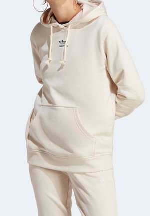 Personne portant un sweat à capuche Adidas beige clair avec poche avant et pantalon assorti, debout devant un fond uni clair.