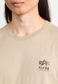 Bež majica s kratkimi rokavi, izdelana iz mehke tkanine, z logotipom, ki vključuje geometrijske oblike in besedilo "ALPHA INDUSTRIES" v temnih barvah.