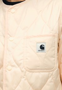 Ανοιχτό ροζ καπιτονέ μπουφάν με κουμπιά τύπου κλικ και τετράγωνη μπροστινή τσέπη που φέρει ένα μαύρο-άσπρο λογότυπο Carhartt.