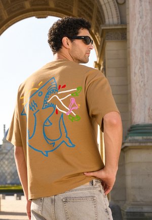 Mann mit Sonnenbrille und beigefarbenem T-Shirt mit buntem Haifisch-Motiv, der Beine mit grünen Schuhen beißt, steht in der Nähe eines steinernen Torbogens im Freien.