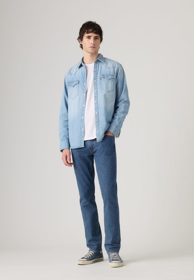 Joven de pie con camisa vaquera azul claro sobre camiseta blanca, vaqueros azules y zapatillas azules sobre fondo liso.