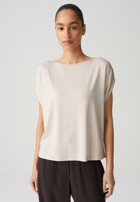 Licht beige t-shirt met korte mouwen en een wijde halslijn, gemaakt van zachte stof. Het heeft een relaxed fit en een gladde textuur. Gecombineerd met donker gestructureerde broek.