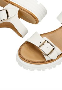 Sandales blanches en cuir avec une large sangle et un détail de boucle dorée, dotées d'une semelle intérieure texturée en tan et d'une épaisse semelle en caoutchouc blanc.