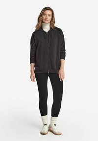 Schwarze Zip-Hoodie mit lockerer Passform und großen Fronttaschen, kombiniert mit schwarzen Leggings und cremefarbenen Socken. Das Model trägt Sneakers.