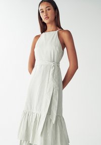 Juostuota suknelė šviesiai žalia ir balta, su špagetinių dirželių petnešomis, raišteliu juosmenyje ir raukšlėta apatine dalimi. Pagaminta iš lengvo audinio.