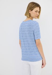 Modré tričko s krátkým rukávem s texturovaným vzorem, kulatým výstřihem a rovného střihu. Nosené s bílými kalhotami, ukazuje neformální design.