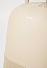 Zaino beige Calvin Klein, parte superiore in pelle liscia, metà inferiore testurizzata con motivo, chiusura con zip superiore, logo stampato in bianco sul davanti.