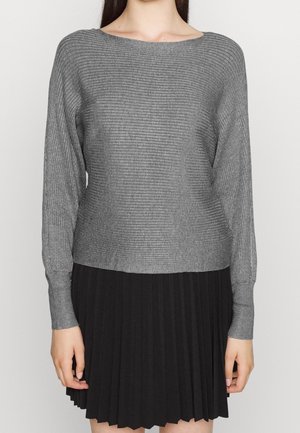 Femme portant un pull gris côtelé à manches longues et une jupe noire plissée, debout devant un arrière-plan clair uni.