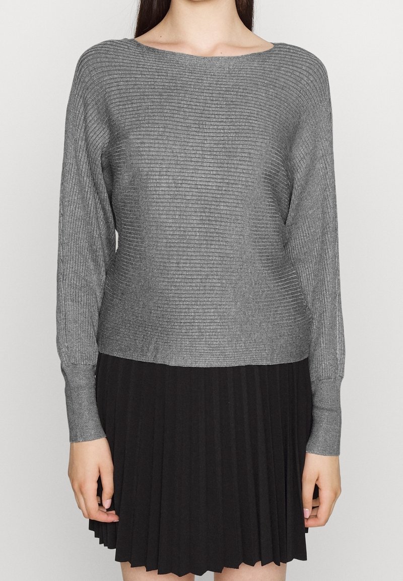 Femme portant un pull gris côtelé à manches longues et une jupe noire plissée, debout devant un arrière-plan clair uni.