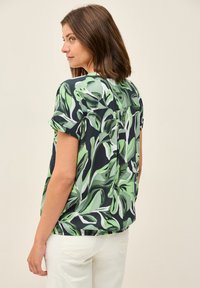 Camicia a maniche corte con un motivo a foglie verdi e nere, realizzata in tessuto leggero con orlo arrotondato e dettaglio di piega sul retro.