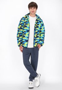 Kamuflage pufferjakke i blå og neon grøn, med hætte og lynlås. Sammen med grå joggers og hvide sneakers.